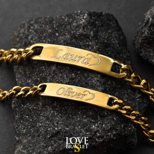 May include: Two gold-colored personalized bracelets with the names "Laura" and "Oliver" engraved on them. Each bracelet has a heart symbol next to the name. The bracelets are on a dark, textured surface. The brand name "LOVE BRASLET" is visible.