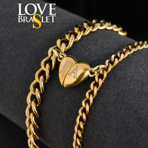 May include: Gold bracelet featuring a heart-shaped pendant split in two, with the letters "J" and "A". The bracelet has a thick chain design. The words "LOVE BRASLET" are in the upper left corner.