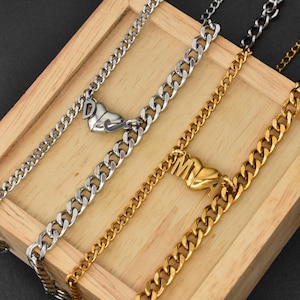 May include: Two curb chain necklaces, one silver-toned and one gold-toned, each featuring a heart-shaped pendant. The silver necklace displays the letter D, while the gold necklace displays the letter M. Both chains have a metallic finish and links.