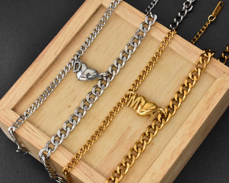 May include: Two curb chain necklaces, one silver-toned and one gold-toned, each featuring a heart-shaped pendant. The silver necklace displays the letter D, while the gold necklace displays the letter M. Both chains have a metallic finish and links.
