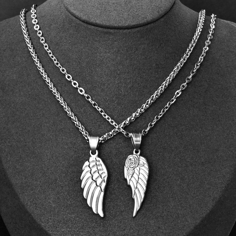 May include: Two silver-colored necklaces with wing pendants. One pendant features a detailed wing design, while the other has a wing with a rose. The necklaces are displayed on a dark gray surface.