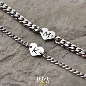 Może przedstawiać: Dwie srebrne bransoletki z łańcuszka z zawieszkami w kształcie serca. Jedna zawieszka ma literę "M", a druga literę "K". Bransoletki leżą na teksturowanej, szarej powierzchni. Słowa "LOVE BRASLET" znajdują się w dolnym rogu.