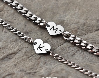 May include: Two silver chain bracelets with heart-shaped charms. One charm has the letter "M" and the other has the letter "K". The bracelets are on a textured, gray surface. The words "LOVE BRASLET" are in the bottom corner.