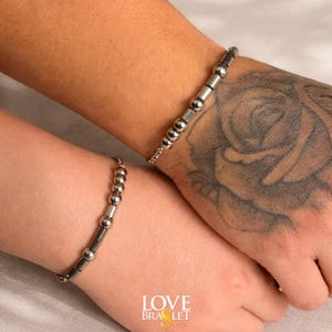 May include: Two silver bracelets with a chain and bead design are shown on wrists. The bracelets have a series of small, round beads and cylindrical segments. The word "LOVE BRASLET" is printed in white and yellow at the bottom of the image.