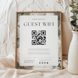 May include: A rectangular gold-toned frame containing a white sign with the text GUEST WIFI, a central QR code, fields for network and password, and social media icons. The sign features a floral border with muted pink and green tones.