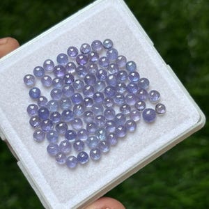 Op de afbeelding: Een verzameling kleine, ronde, gepolijste edelstenen in tinten blauw en paars. De edelstenen zitten in een witte, vierkante plastic bak. De edelstenen hebben een glad, reflecterend oppervlak.