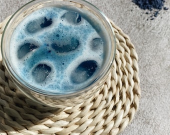 May include: A glass of blue iced beverage with foam on top, sitting on a woven coaster. A small pile of blue powder is visible in the background.