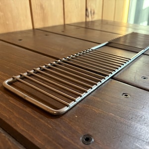 May include: A stainless steel grill grate with parallel bars and a rectangular shape. The grate is designed for cooking and is placed on a dark brown wooden surface. The metal has a shiny, reflective quality.