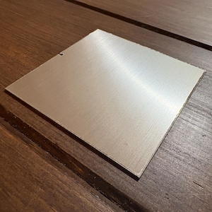 May include: A square, brushed metal plate with a silver finish. The plate has a slightly rough edge and is set against a dark brown wooden surface. The metal surface reflects light, highlighting the brushed texture.