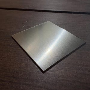 May include: A square, brushed metal plate with a reflective surface. The plate is positioned on a dark wooden surface, showcasing its metallic sheen. The edges are clean and defined, highlighting the plate's geometric shape.