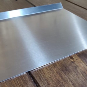 May include: A rectangular, stainless steel metal spatula with a raised edge. The spatula has a brushed finish and is set on a wooden surface.