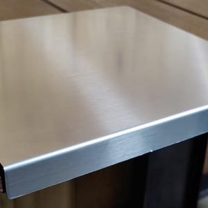 May include: A stainless steel table top protector with a brushed finish. The square-shaped protector has a slightly raised edge and is placed on a wooden table. The wood has a warm, brown tone.