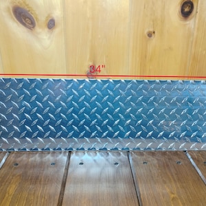May include: A rectangular, diamond-plate metal sheet, 84 inches long and 8 inches wide. The surface has a repeating pattern of raised, diamond-shaped treads. The metal has a shiny, silver appearance, reflecting light. The sheet is displayed on a wooden surface.