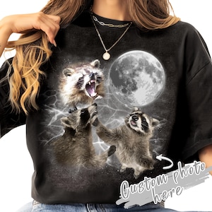 May include: Black t-shirt with a graphic of three raccoons under a full moon and lightning. One raccoon is roaring, while the others are looking up. The text "Custom photo here" is also visible.