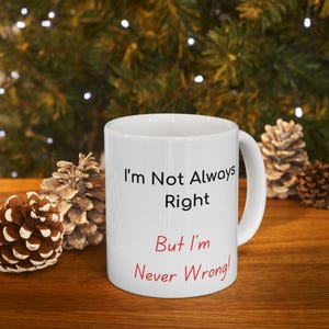 May include: White ceramic mug with the text "I'm Not Always Right But I'm Never Wrong!" in black and red. The mug is on a wooden surface with pine cones and a blurred green background.