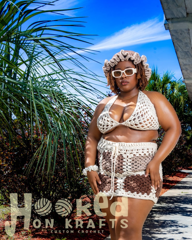 May include: A woman wearing a white crochet bikini top and skirt with a brown and white pattern. She is wearing a white crocheted hat and sunglasses. The text "Hooked On Krafts Custom Crochet" is visible at the bottom of the image.