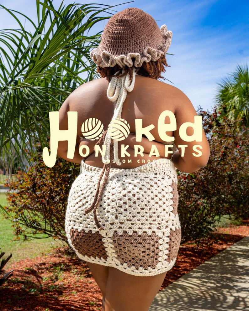 May include: A crocheted mini dress in brown and white, featuring a halter top and a skirt with a geometric pattern. The model is wearing a brown crocheted hat with a ruffled brim. The text "Hooked On Krafts Custom Crochet" is visible on the dress.