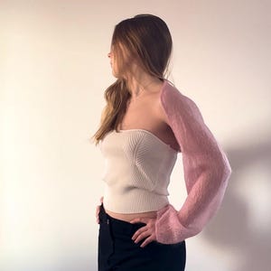 Könnte beinhalten: Eine Frau trägt ein weißes Crop-Top und eine schwarze Hose mit einer rosafarbenen Strickjacke über den Schultern.