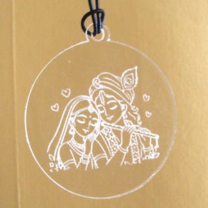 May include: A circular clear acrylic pendant featuring a white laser-etched illustration of Radha and Krishna. The design shows Krishna playing a flute beside Radha, surrounded by small heart outlines. A black cord is looped through the top hole.
