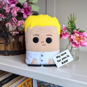 May include: A plush toy of a person with yellow hair, a blue shirt, and a smiling face. The toy is holding a sign that says "My love for you is RAW".
