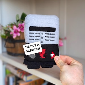 May include: A soft toy depicting a knight's helmet and torso, with a grey helmet, black tunic, and red dragon embroidery. The text "TIS BUT A SCRATCH" is on a white tag. The toy is held by a hand.