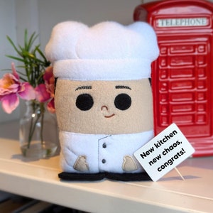 May include: A plush toy chef with a white chef hat, black hair, and a brown face. The chef is wearing a white chef's uniform with black pants. The toy is holding a sign that says "New kitchen, new chaos, congrats!"