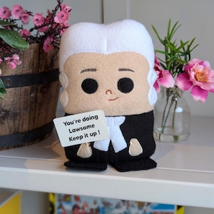 May include: A plush toy of a judge wearing a black robe and white wig. The toy is holding a sign that says "You're doing Lawsome Keep it up!"