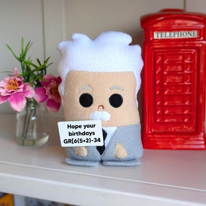 May include: A handmade felt doll resembling Albert Einstein, with white hair, a mustache, and a gray suit. The doll holds a sign that reads "Hope your birthdays GR[6(5+2)-34". A red telephone booth and pink flowers in a glass vase are in the background.