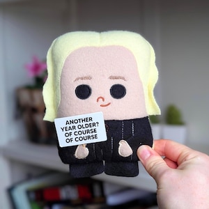 May include: A felt doll with a cartoon design, featuring blonde hair and black clothing. The doll holds a small white sign that reads, "ANOTHER YEAR OLDER? OF COURSE OF COURSE."