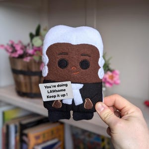 May include: A felt doll resembling a judge, featuring a white wig, brown skin, and a black robe. The doll holds a sign with the text "You're doing LAWsome Keep it up!" The doll has a smiling face and is held by a hand.