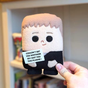 May include: A rectangular plush toy with a light brown hairstyle and black eyes, dressed in a black suit. It holds a white sign with the text: "COULDN'T GET YOU WHITAKER SO I GOT YOU THIS PLUSHIE!"
