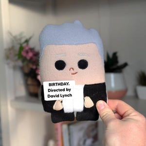 May include: A handmade plush doll with a light blue hair, black eyes, and a small smile. The doll is wearing a black suit and holding a white sign that reads "BIRTHDAY. Directed by David Lynch."