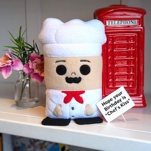 May include: A plush chef doll wearing a white chef's hat and a red bow tie. The doll has a brown face with black eyes, a black moustache, and a white smile. The doll is standing on a white shelf with a red telephone box behind it. A white card with black text reads "Hope your Birthday is *Chef's Kiss*"