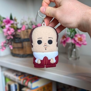 May include: A felt keychain depicting a stylized figure with a light skin tone, dark hair, and a small goatee. The figure wears a white collar and a burgundy outfit. The keychain has a silver metal ring attached to the top.