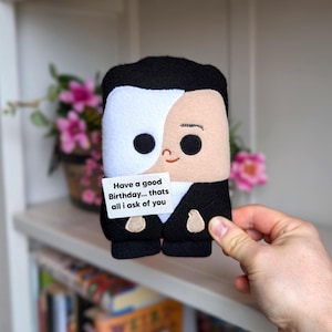 Peut inclure: Poupée en peluche d'un personnage avec un masque noir et blanc, portant un costume noir. La poupée tient une petite carte blanche avec le texte "Have a good Birthday... thats all i ask of you". La poupée est tenue devant une étagère avec des fleurs.