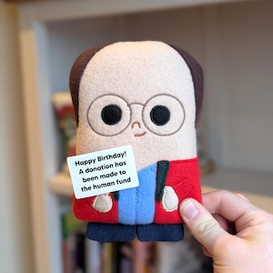 May include: A plush doll with a light skin tone, brown hair, and round glasses. The doll wears a red jacket, blue shirt, and black pants. A white card reads "Happy Birthday! A donation has been made to the human fund."