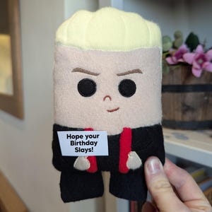 May include: A plush toy shaped like a person with blonde hair, wearing a black suit with a red tie. The toy has a message on a white tag that says "Hope your Birthday Slays!"