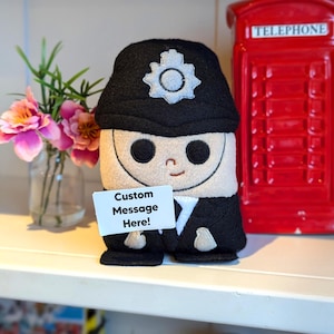 May include: A plush toy of a British police officer wearing a black hat with a silver badge. The toy is holding a white sign that says "Custom Message Here!"