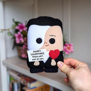 May include: A plush doll of a character with a white mask and black hair, holding a red heart and a sign that reads "Happy Valentines, Thats all I ask of you." The doll is wearing a black tuxedo.