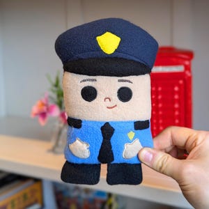 May include: A plush toy police officer with a blue hat, blue uniform, and a black tie. The toy has a beige face with embroidered eyes, eyebrows, and a smile.