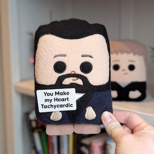 May include: A handmade plush doll with a black beard and hair, wearing a navy blue jacket, and holding a sign that reads "You Make my Heart Tachycardic." Another doll is visible in the background. The dolls are made of felt.