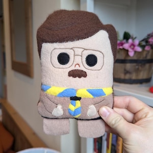 May include: A brown plush toy with a yellow and blue scarf, brown hair, glasses, and a mustache. The toy is holding its hands in front of it.