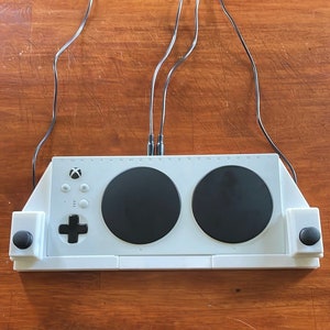 May include: White gaming controller with two round black buttons and a white base. The controller has a black Xbox logo and a black directional pad. The controller is connected to a white surface with two black cables.