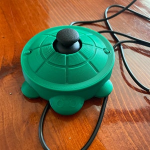May include: A green and black joystick with a round base and a black knob. The joystick is connected to a black cord.
