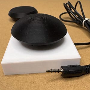 May include: A black, dome-shaped button with a textured surface sits atop a white, rectangular base. A black cable extends from the base, terminating in a standard 3.5mm audio jack connector. A second, smaller black dome-shaped button is visible nearby.