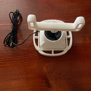 May include: White 3D printed phone stand with a rotating base and a black cord.