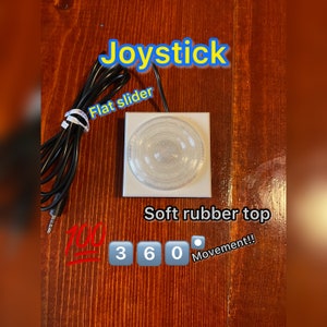 May include: A white square joystick with a soft rubber top and a flat slider. The joystick has a 360 degree movement and is connected to a device with a black cord and a 3.5mm jack. The text "Joystick" is written in yellow above the joystick, and the text "Soft rubber top" is written below the joystick. The text "360 Movement!" is written below the joystick.