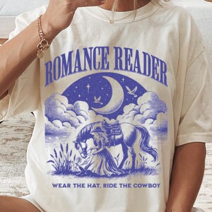 May include: A cream colored t-shirt with a purple graphic of a woman riding a horse under a crescent moon and stars. The text "Romance Reader" and "Wear the hat, ride the cowboy" is printed on the shirt.
