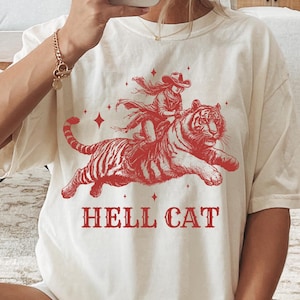 Könnte beinhalten: Ein weißes T-Shirt mit einem roten Aufdruck einer Frau, die auf einem Tiger reitet. Der Aufdruck enthält Sterne und den Text "HELL CAT".