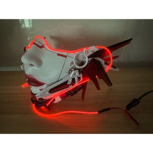 Może przedstawiać: Biała i czerwona plastikowa maska z czerwonym świecącym światłem. Maska ma ludzką twarz z detalicznym wzorem.